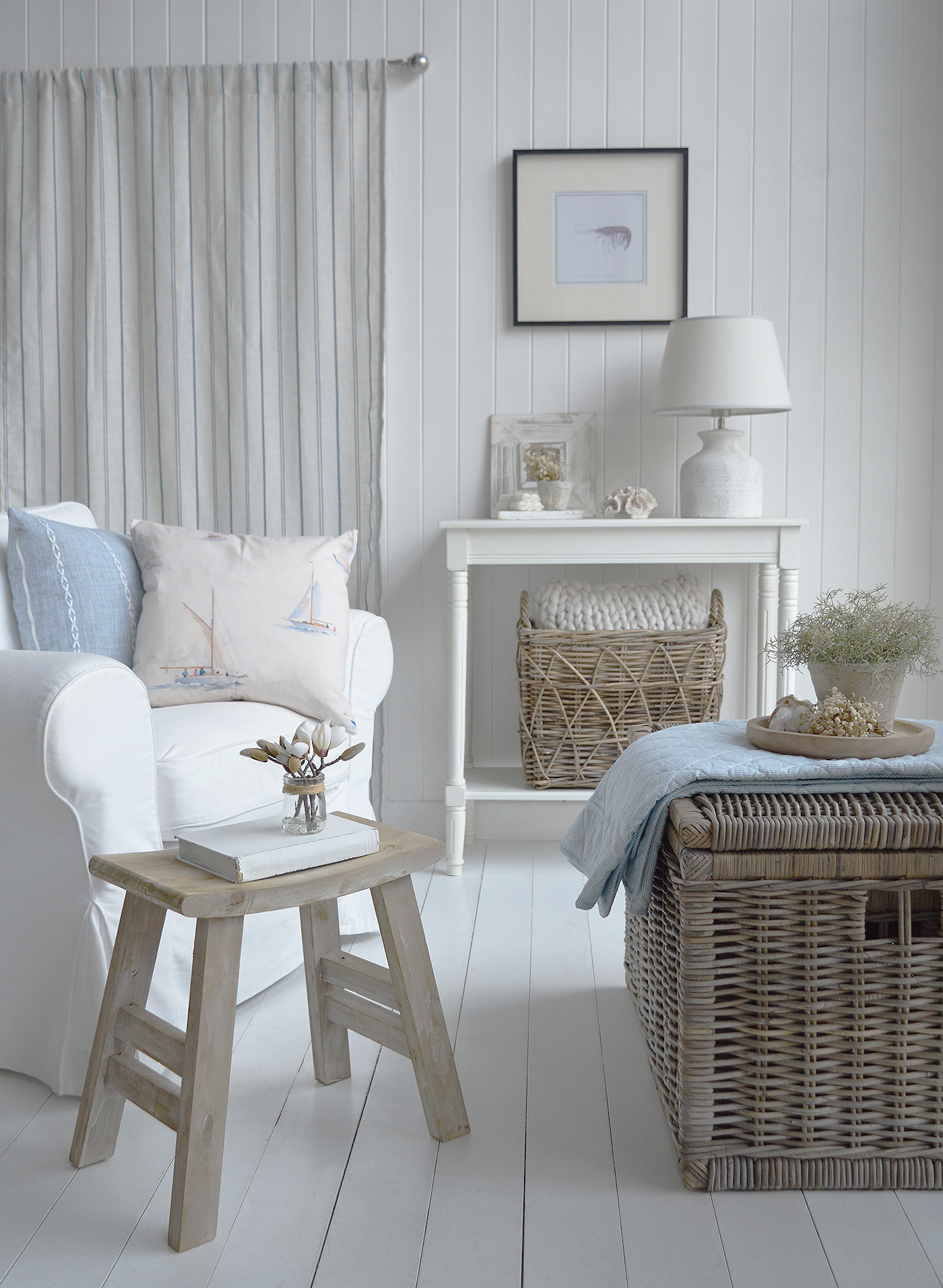 A calm, coastal living space styled with white shiplap walls, a neutral linen armchair, soft blue nautical cushions, woven rattan storage, and light-washed wooden furniture, creating a relaxed Hamptons-inspired coastal interior with a fresh, airy feel.