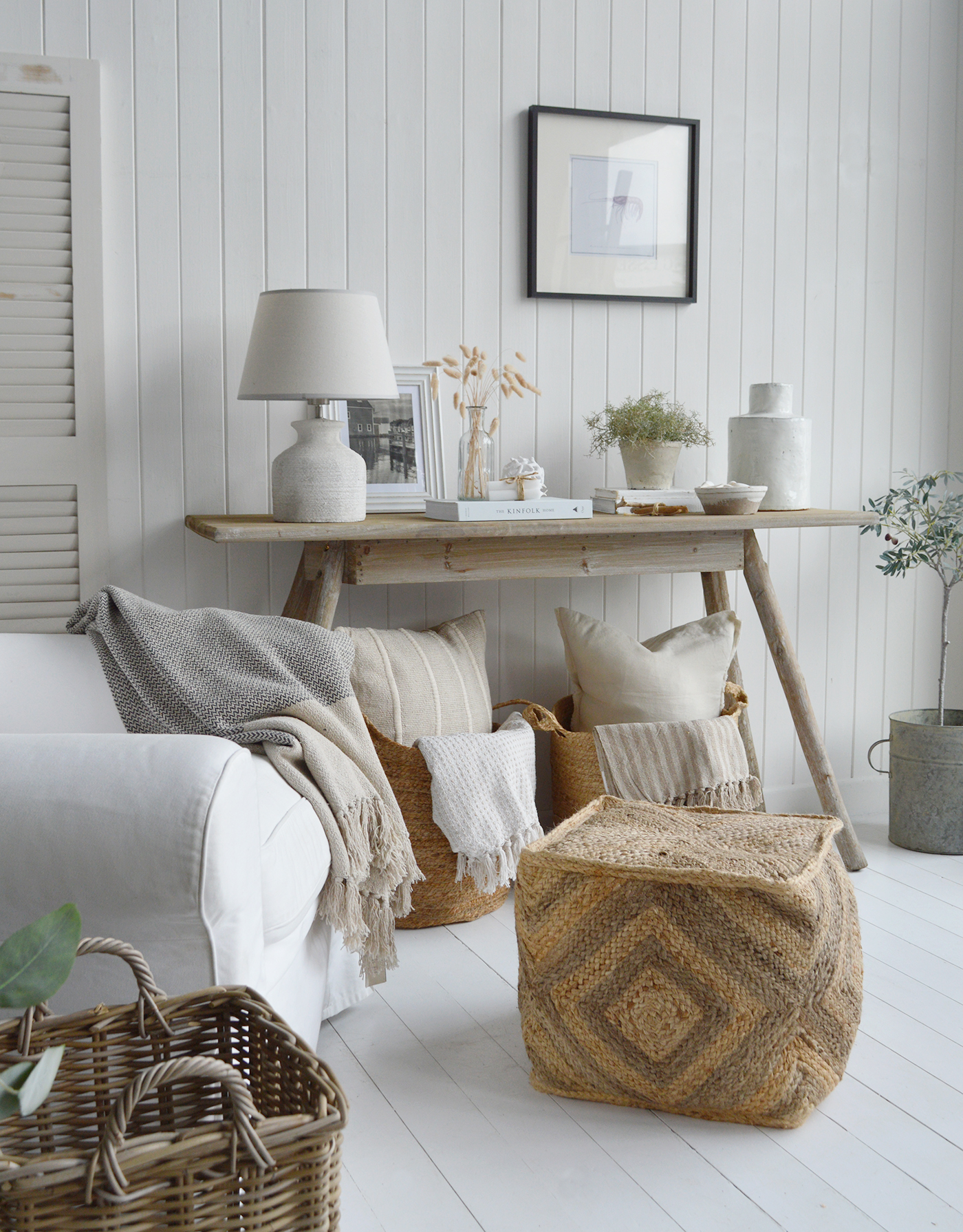 Modern coastal interiors with rustic wood console table, ceramic lamp, framed art, neutral pillows and throws in woven baskets, jute pouf, wicker basket, white slipcovered sofa, and bright white paneled walls for relaxed beach-inspired home decor